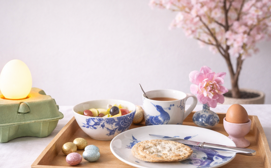 Minimalistisch paasontbijt op wit tafelkleed met eivormige lamp in groene eierdoos, houten dienblad met Delfts blauw servies, broodje en thee, met roze bloesem op de achtergrond.