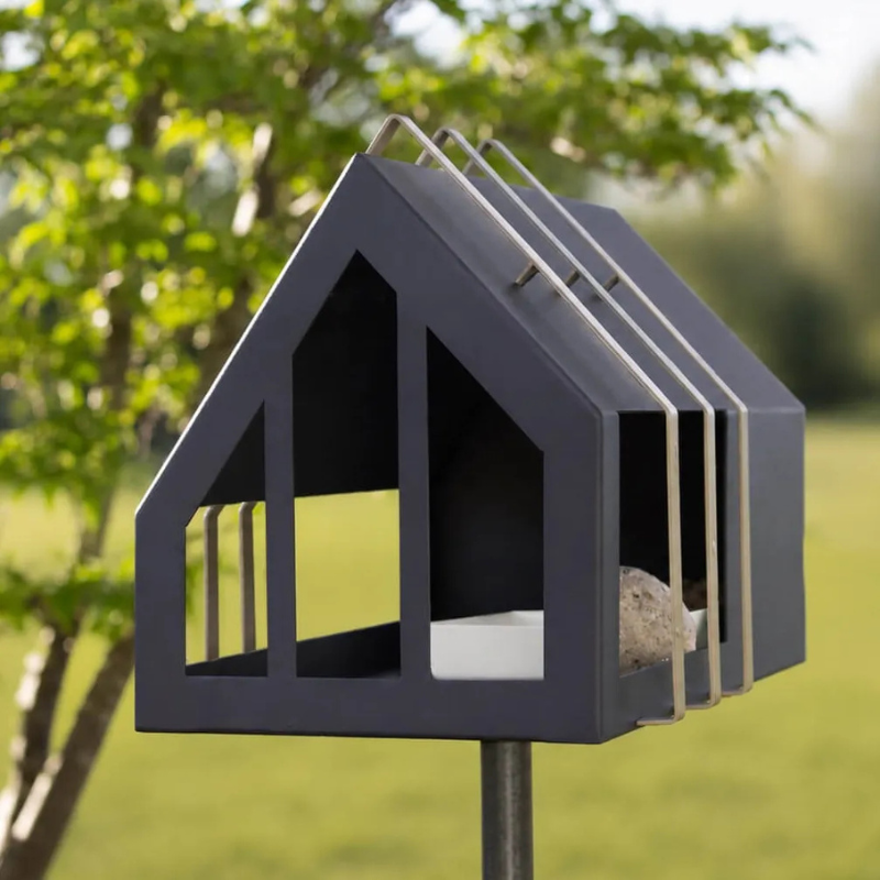 Metalen vogelvoederhuis op paal in de tuin met een mees bij het witte voederschaaltje